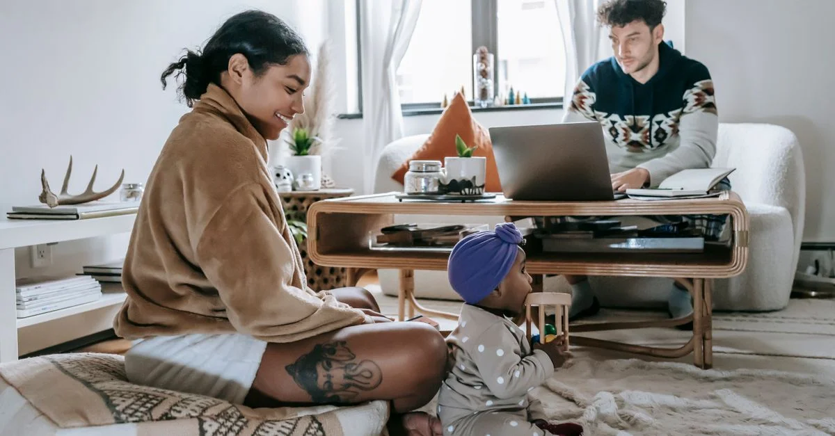 Mère et bébé nouant des liens dans un salon confortable pendant que le père travaille à distance sur un ordinateur portable.
