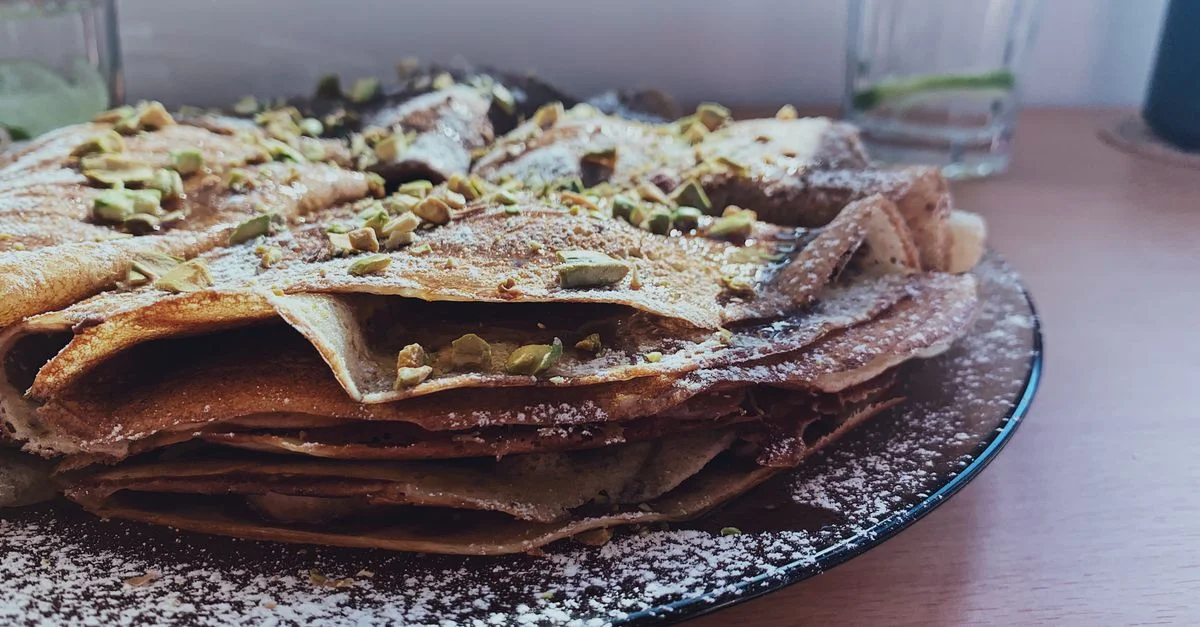 Vue rapprochée d'une pile de crêpes maison garnies de sucre glace et de pistaches sur une table.
