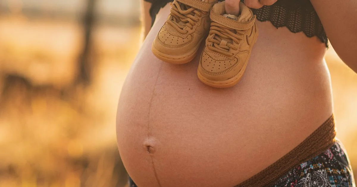 Mujer embarazada al aire libre sosteniendo zapatos de bebé sobre su barriga, simbolizando la maternidad.
