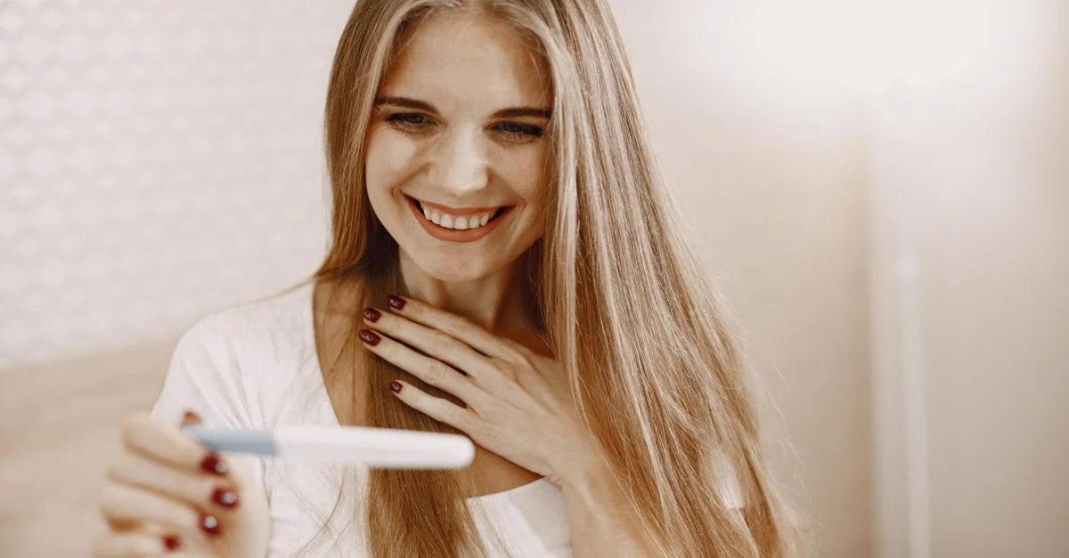 Mujer sonriente vestida de blanco con cabello largo encantada por el resultado positivo de la prueba de embarazo en interiores.