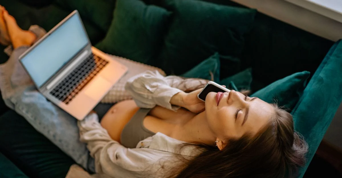 Una mujer embarazada descansando en un sofá, con los ojos cerrados, hablando por un smartphone con una laptop cercana.