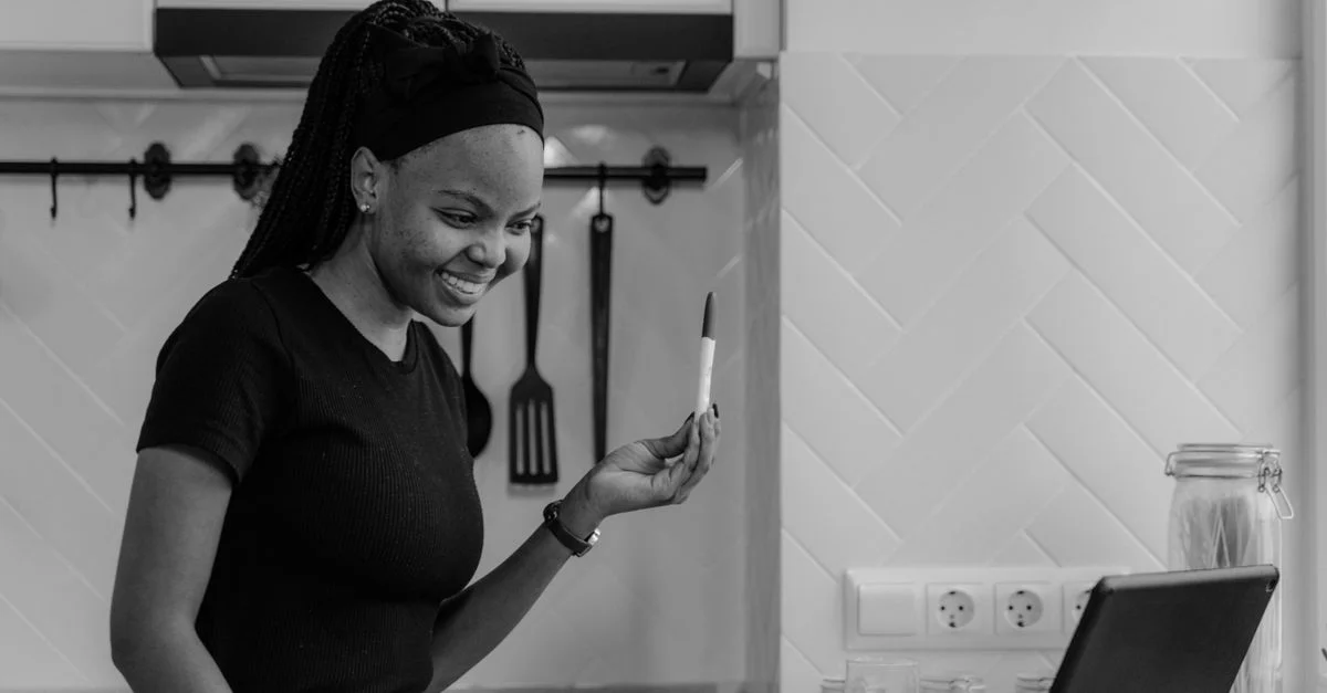 Una mujer feliz sonriendo a un test de embarazo positivo durante una videollamada en una cocina moderna.