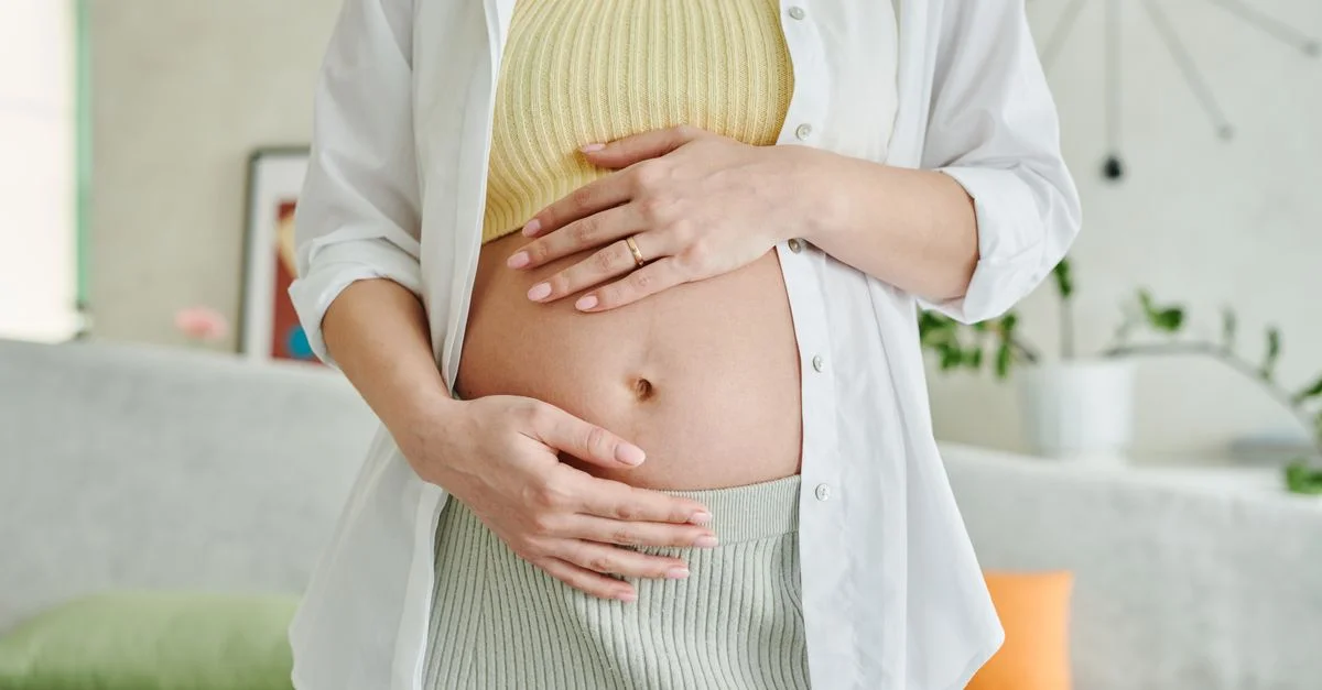Una imagen en primer plano de una mujer embarazada sosteniendo suavemente su barriga en interiores.