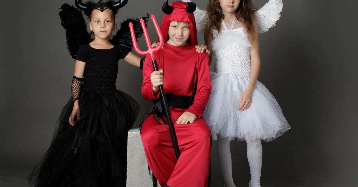 Tres niños disfrazados de diablo y ángeles posando en interiores para una foto temática de Halloween.