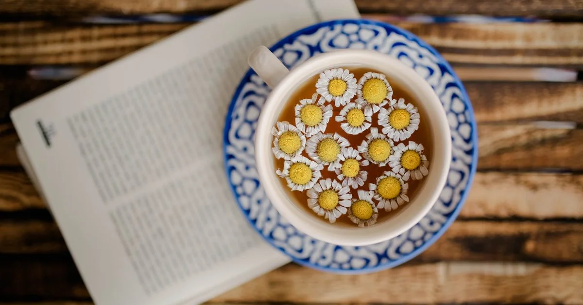 Toma aérea de té de manzanilla en una taza decorativa con un libro abierto, transmitiendo una vibra tranquila y acogedora.