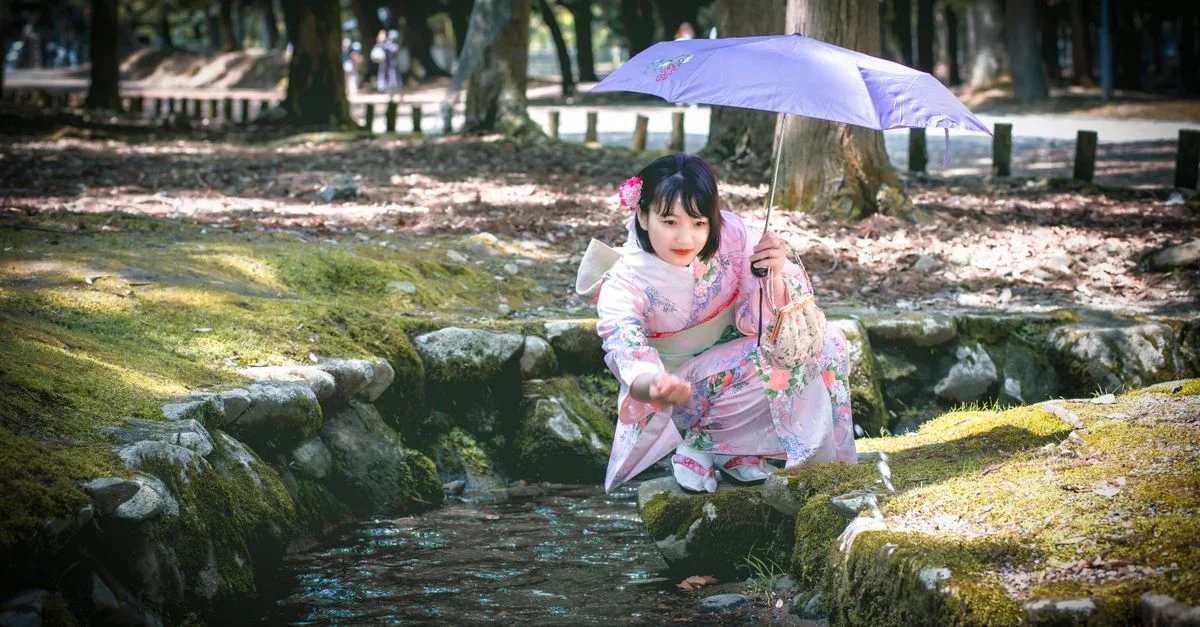 Una niña con un kimono floral sosteniendo un parasol junto a un arroyo sereno en un entorno japonés tradicional.