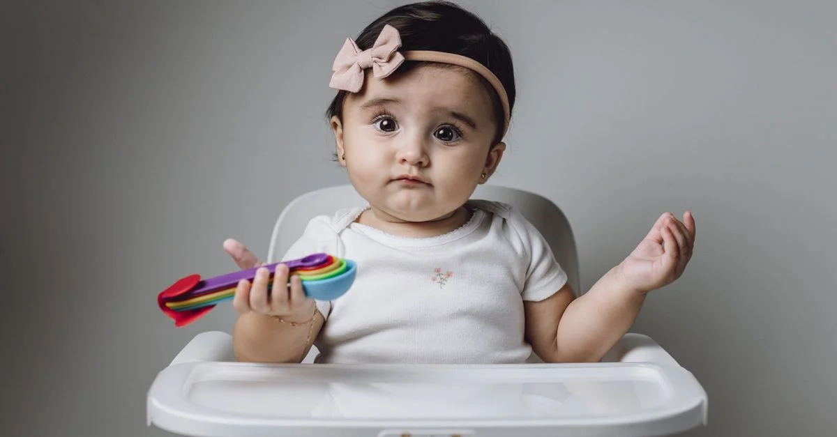 Linda niña en una trona sosteniendo una cuchara colorida, en un entorno interior simple.
