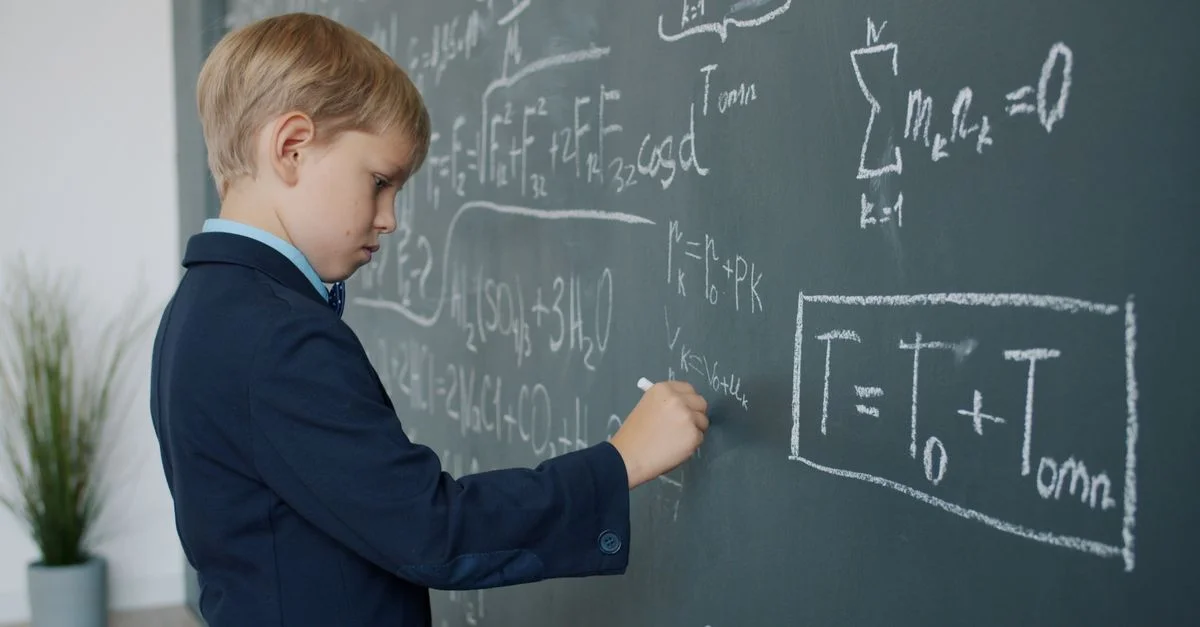 Un niño con traje escribe ecuaciones matemáticas complejas en la pizarra de un aula.