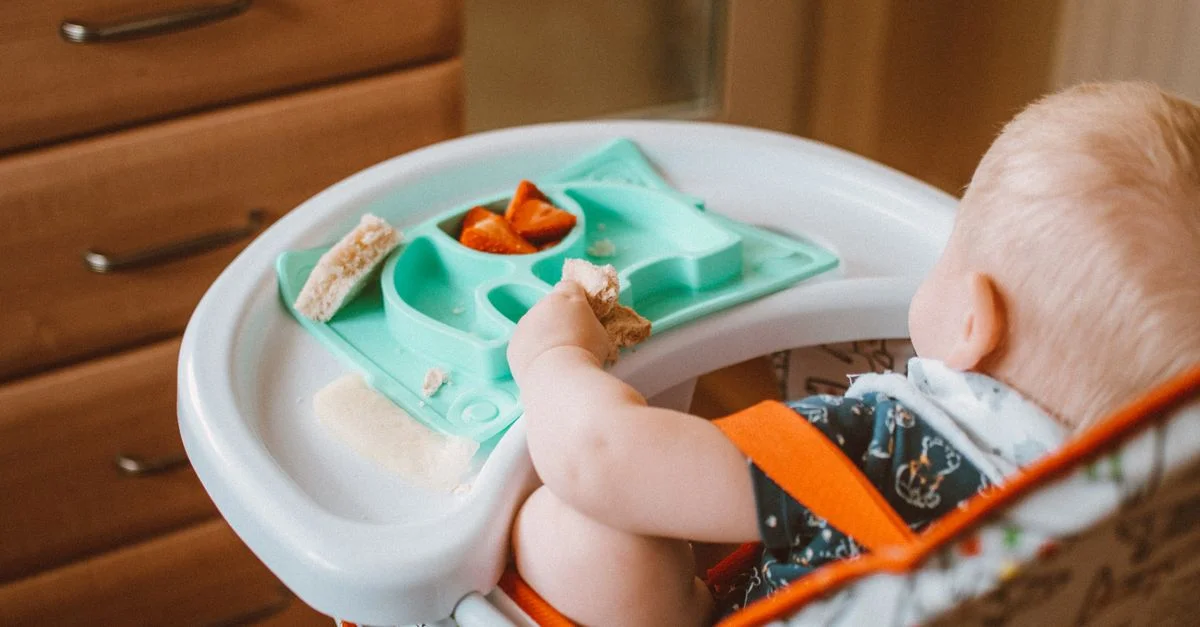 Un lindo bebé disfrutando de la hora del snack en una trona con bandeja de comida colorida en interiores.
