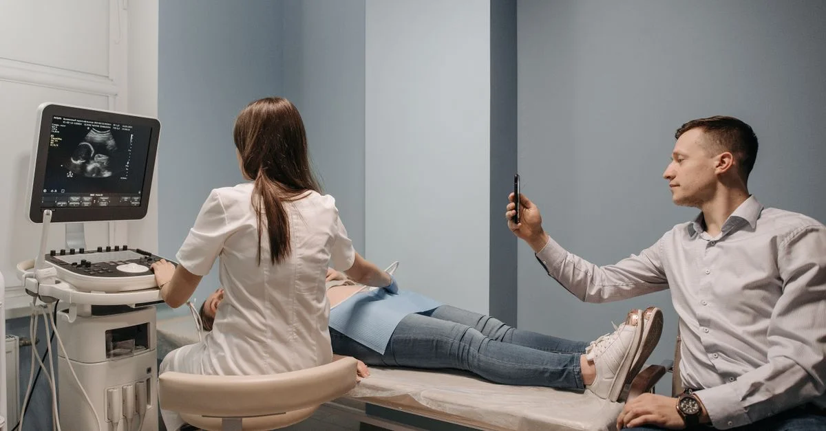 Un médico realiza una ecografía a una mujer embarazada, mientras un hombre toma una foto en un entorno clínico.