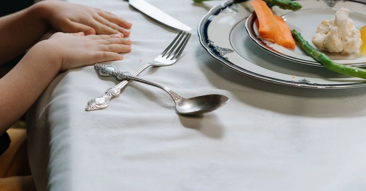 Las manos de un niño en una mesa de comedor con platos de verduras, mostrando una comida saludable.