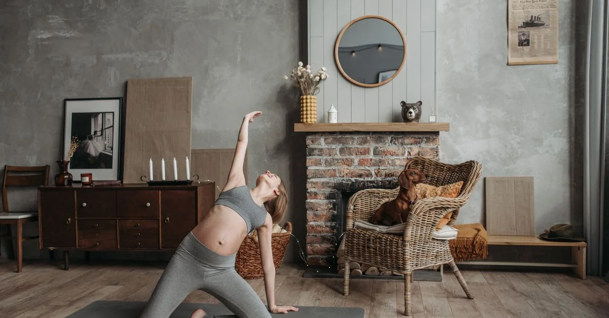 Mujer embarazada practicando yoga cerca de una chimenea con un perro en una habitación acogedora.